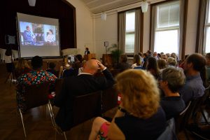 Ein Saal mit vielen sitzenden Menschen. Vorne wird auf einer Leinwand ein Film abgespielt.