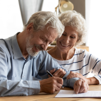 Ein älteres Paaar (mann und Frau) sitzen an einem Tisch. Der Mann schreibt etwas auf einen Papierblock.