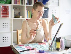 eine Frau mit kurzen Haaren hält eine Tube Farbe in der einen und einen Pinsel in der anderen Hand.